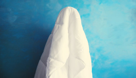 Cheerful lazy woman waking up after sleeping covered head with white blanket on blue background at homeの写真素材