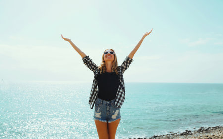 Summer vacation, happy smiling woman raising her hands up on the beach on sea coast backgroundの写真素材