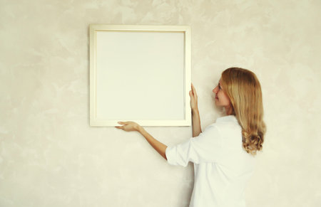 Young woman decorating interior, hanging blank a photo frame mockup on white wall in new home