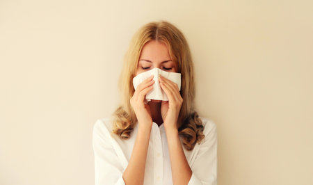 Sick exhausted caucasian young woman employee sneezing blow nose using tissue at homeの写真素材
