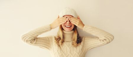 Portrait of cheerful laughing happy young woman covering her eyes with hands in warm soft knitted clothes, sweater and hat on beige backgroundの写真素材