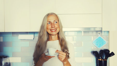 Portrait of happy smiling middle-aged gray-haired woman enjoying hot tasty cup of coffee or tea in the kitchen in early morning at homeの写真素材