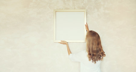 Young woman decorating interior, hanging blank a photo frame mockup on white wall in new homeの写真素材