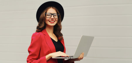 Portrait of businesswoman working with laptop wearing red blazer jacket, eyeglasses, round hat on gray background, happy modern young woman holds pc computerの写真素材