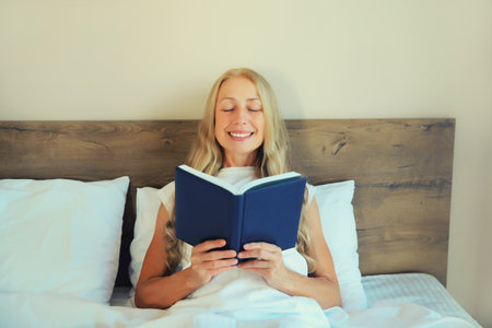 Happy middle aged woman reading book with blank cover while lying on the bed at homeの写真素材