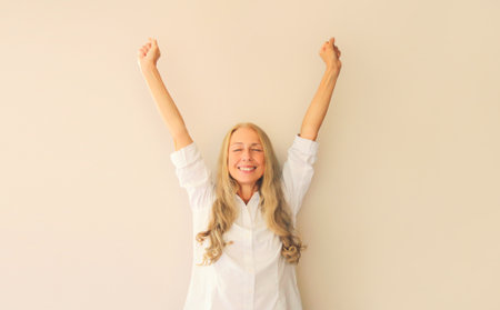 Happy lucky caucasian middle-aged woman employee manager in white office shirt having fun or feels victory stretching or raising her hands up at work or homeの写真素材