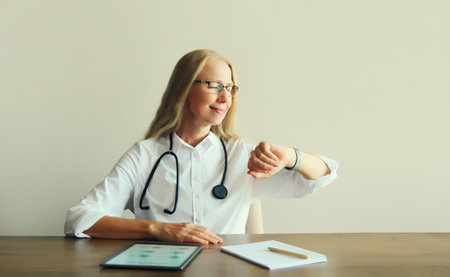 Caucasian middle-aged woman doctor with stethoscope, paper documents and digital tablet computer sitting at desk in clinic officeの写真素材