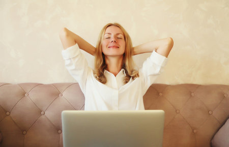 Relaxed caucasian young woman taking a break while working with laptop sitting on couch in room at homeの写真素材