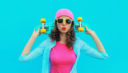 Portrait of stylish modern young woman with skateboard posing in colorful vivid clothes, pink hat on blue studio backgroundの写真素材