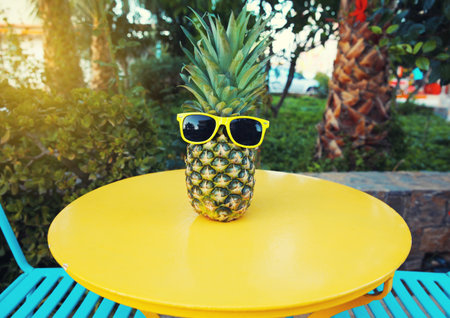 Summer image of stylish pineapple fruit in yellow sunglasses on the table in street cafe in city...