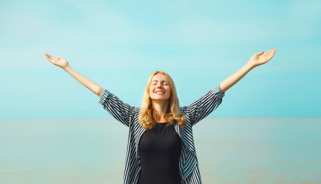Summer vacation, happy young woman raising her hands up on the beach on sea coast and blue sky...