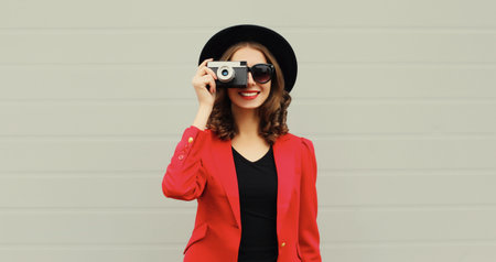 Beautiful stylish young woman photographer with film camera taking picture in black round hat, red...