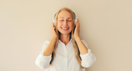 Portrait of happy relaxed middle aged woman listening to music in wireless headphones on white...