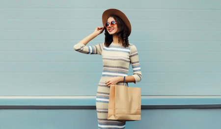 Stylish beautiful happy smiling young woman posing with shopping bags in round hat, dress on city streetの写真素材