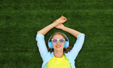 Portrait of happy smiling young woman listening to music in headphones while lying on grass in...