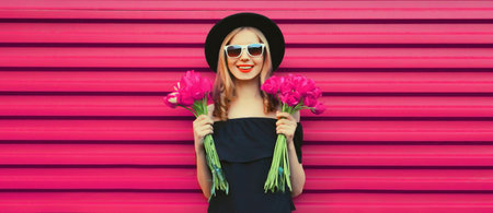 Sweet portrait of beautiful happy smiling woman with bouquet of pink rose flowers posing in black round hat on colorful backgroundの写真素材