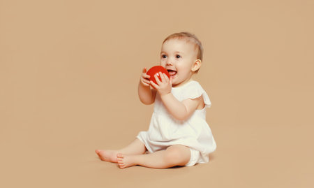 Portrait of cute baby holding red apple fruit sitting on the floor on brown studio backgroundの写真素材
