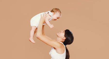 Happy cheerful smiling young mother playing with cute baby on brown studio backgroundの写真素材