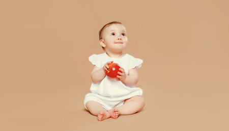 Portrait of cute baby holding red apple fruit sitting on the floor on brown studio backgroundの写真素材