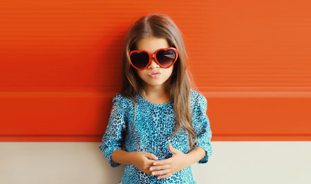 Portrait of beautiful little girl child in red heart shaped sunglasses and blue dress on city streetの写真素材