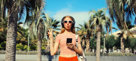 Happy smiling young woman listening to music in headphones with smartphone in summer park against a...