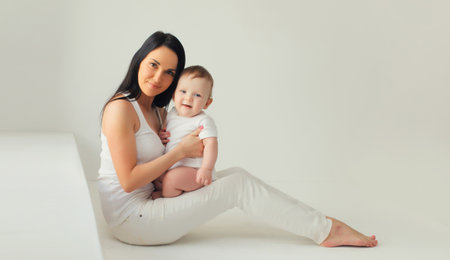 Happy young mother holding her cute baby sitting together in white room at homeの写真素材