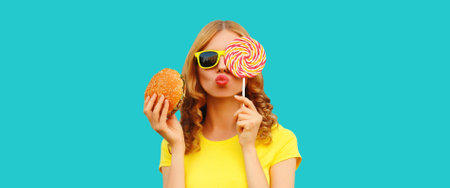 Portrait of happy cheerful young woman having fun with burger fast food and colorful sweet lollipop...