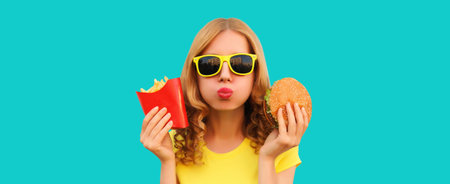 Portrait of happy cheerful young woman eating burger fast food and french fries, fried potatoes isolated on blue studio backgroundの写真素材