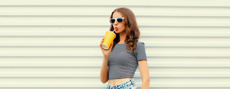 Stylish young woman drinking fresh juice wearing glasses looking away on white wall backgroundの写真素材