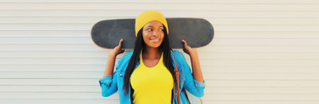 Happy smiling young african woman in colorful yellow hat posing with skateboard in the city on white wall backgroundの写真素材