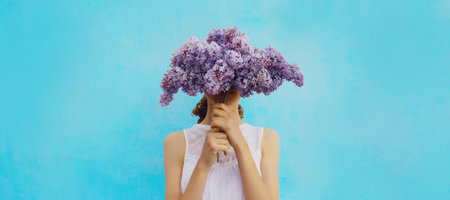 Portrait of woman covering her head with bouquet of fresh lilac flowers on blue backgroundの写真素材