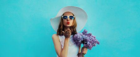 Summer portrait of beautiful lovely young woman blowing kiss with bouquet of purple lilac flowers in white straw hat on blue backgroundの写真素材