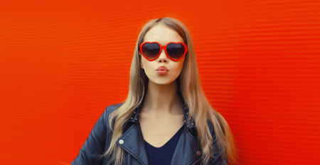 Portrait of beautiful stylish blonde young woman model blowing kiss in red heart shaped sunglasses,...