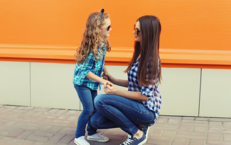 Beautiful stylish mother and child daughter together, happy mom with kid girl looking at each other in glasses, casual clothing in the cityの写真素材