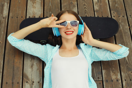 Happy young woman listening to music with headphones lying on a skateboard, modern teenager girl resting in city parkの写真素材