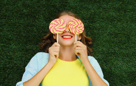 Portrait of happy young woman holding colorful lollipop, cheerful teenager girl lying on green grass in summer park, top viewの写真素材