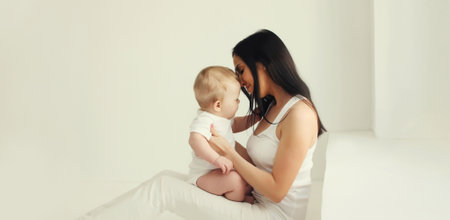 Happy young mother holding her cute baby sitting together in white room at homeの写真素材