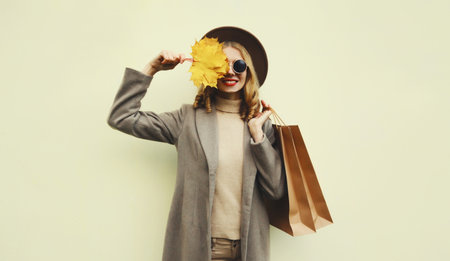 Autumn portrait of beautiful happy young woman with shopping bag, stylish girl holding yellow maple leaves in round hat, coat on brown studio backgroundの写真素材