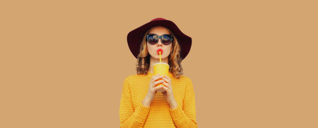 Portrait of stylish young woman drinking fresh juice or coffee, fashionable girl in hat, yellow sweater on brown backgroundの写真素材