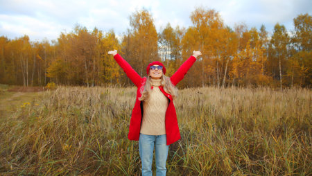 Happy smiling woman in autumn park raising her hands up, stylish joyful girl enjoying warm weather...