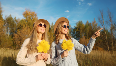 Happy young two women friends with autumn yellow leaves, stylish smiling girlfriends looking away pointing her finger to the side outdoors, in sunny park, fall seasonの写真素材