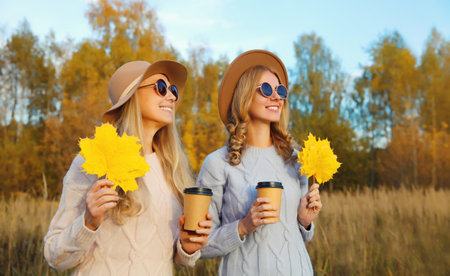 Portrait of happy young two women friends with autumn yellow leaves, stylish smiling girlfriends...