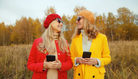 Portrait happy young two women friends talking with phone in autumn park, stylish smiling girlfriends holding smartphone standing together in red yellow coat jacket, hat, outdoors, warm fall seasonの写真素材