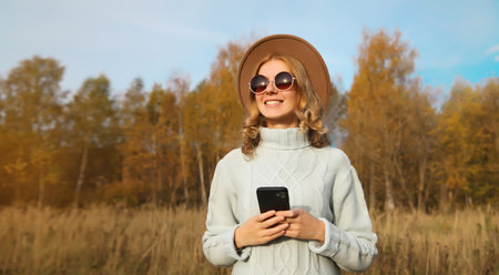 Stylish happy smiling modern young woman with phone standing in autumn park, girl holding smartphone...