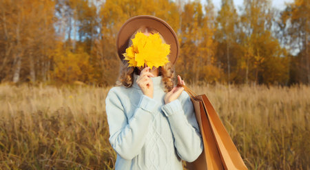 Autumn portrait of stylish happy young woman with shopping bag and maple yellow leaves, fashionable girl smiling wearing glasses, hat, sweater outdoors in park, warm fall seasonの写真素材
