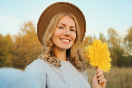 Close up portrait of happy smiling young woman taking selfie with smartphone, yellow leaves in autumn park, joyful girl in hat outdoors, fall seasonの写真素材