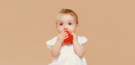 Portrait of cute baby holding red apple fruit sitting on the floor on brown studio background