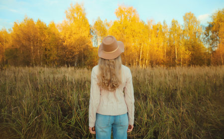 Back view of beautiful happy young woman in brown hat enjoying warm weather, looking at landscape in autumn park with yellow leaves outdoors, fall season, nature backgroundの写真素材