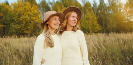 Happy two women friends looking away in autumn park, stylish smiling girlfriends standing outdoors in hat, white sweater together, trees with yellow leaves, nature backgroundの写真素材