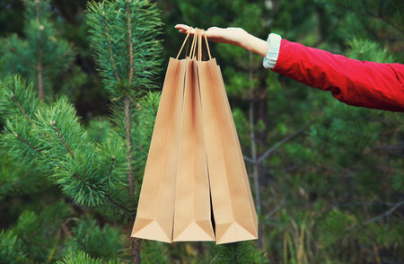 Purchase, holidays, female hand holding showing paper shopping bags, gift against christmas tree...